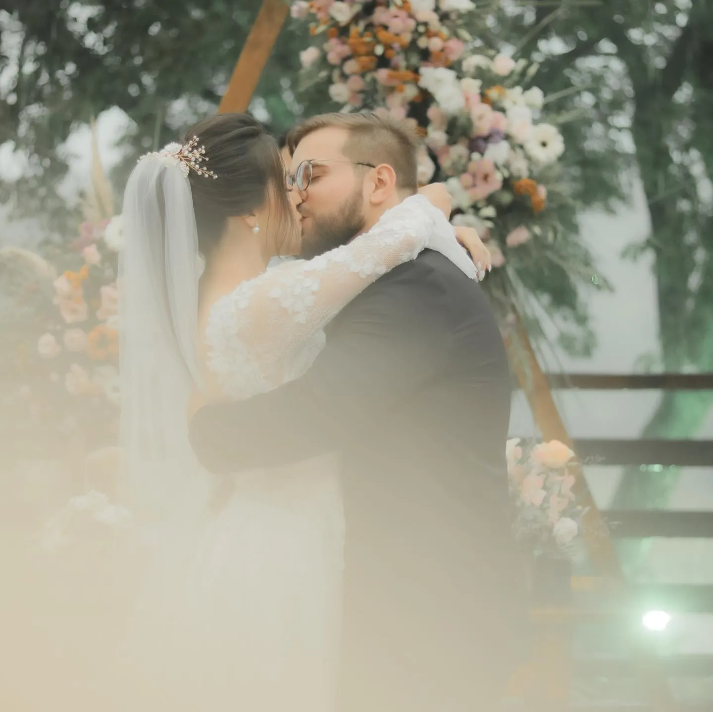 Beijo dos noivos sob o arco de flores, envolto em luz suave
